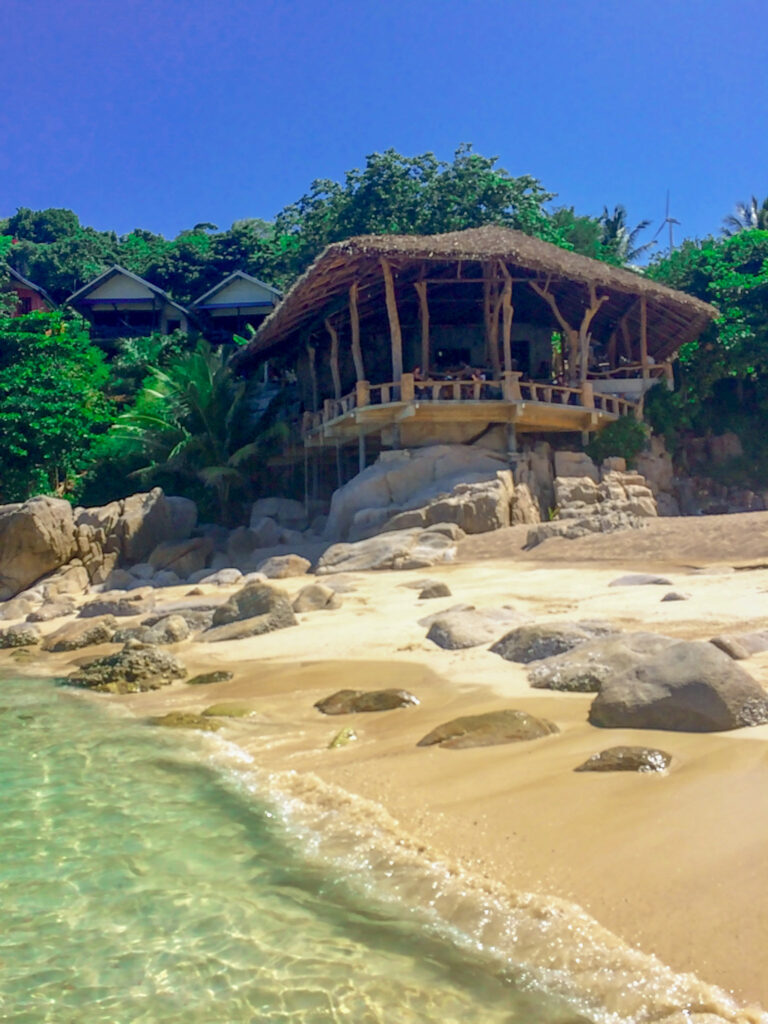 Showcases a beautiful view of the beach, one of the best things to do in Koh Tao Thailand. The white sand beach is set against the crashing waves of the turquoise water in the Gulf of Thailand. A beachfront bar with ocean views is featured.